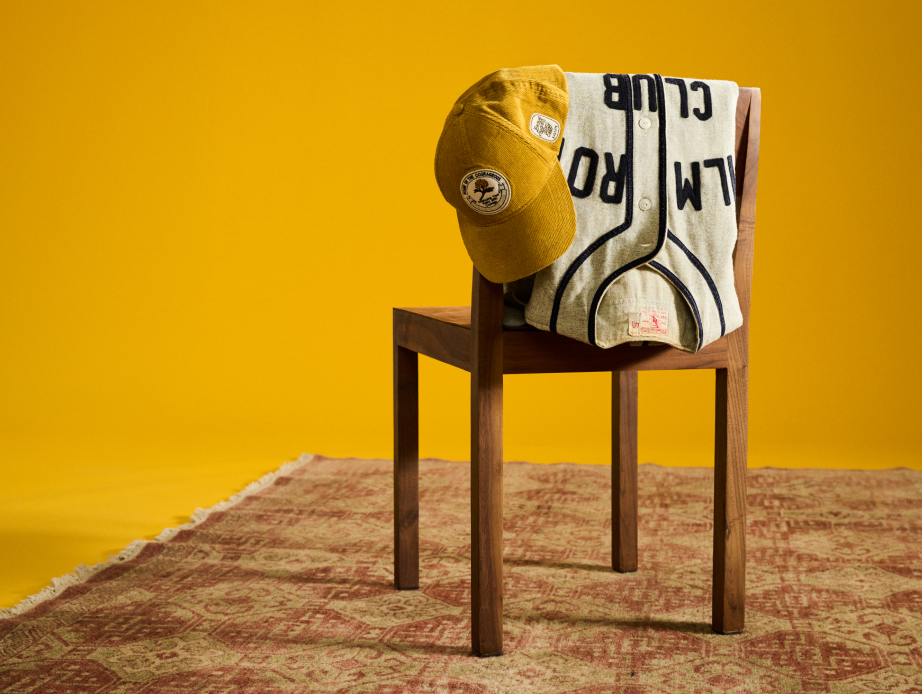A yellow baseball cap and white baseball jersey draped over a wooden chair on a warm-toned rug, against a deep yellow background